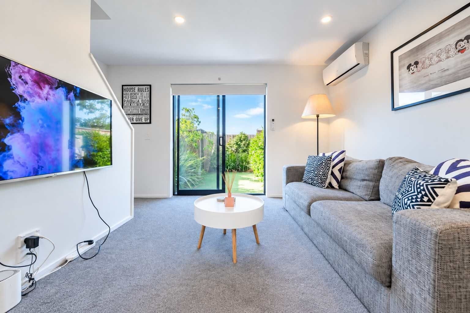 A modern living room with a flat-screen TV, a round coffee table, a grey sofa with patterned cushions, and a large window offering a view of the outdoors.