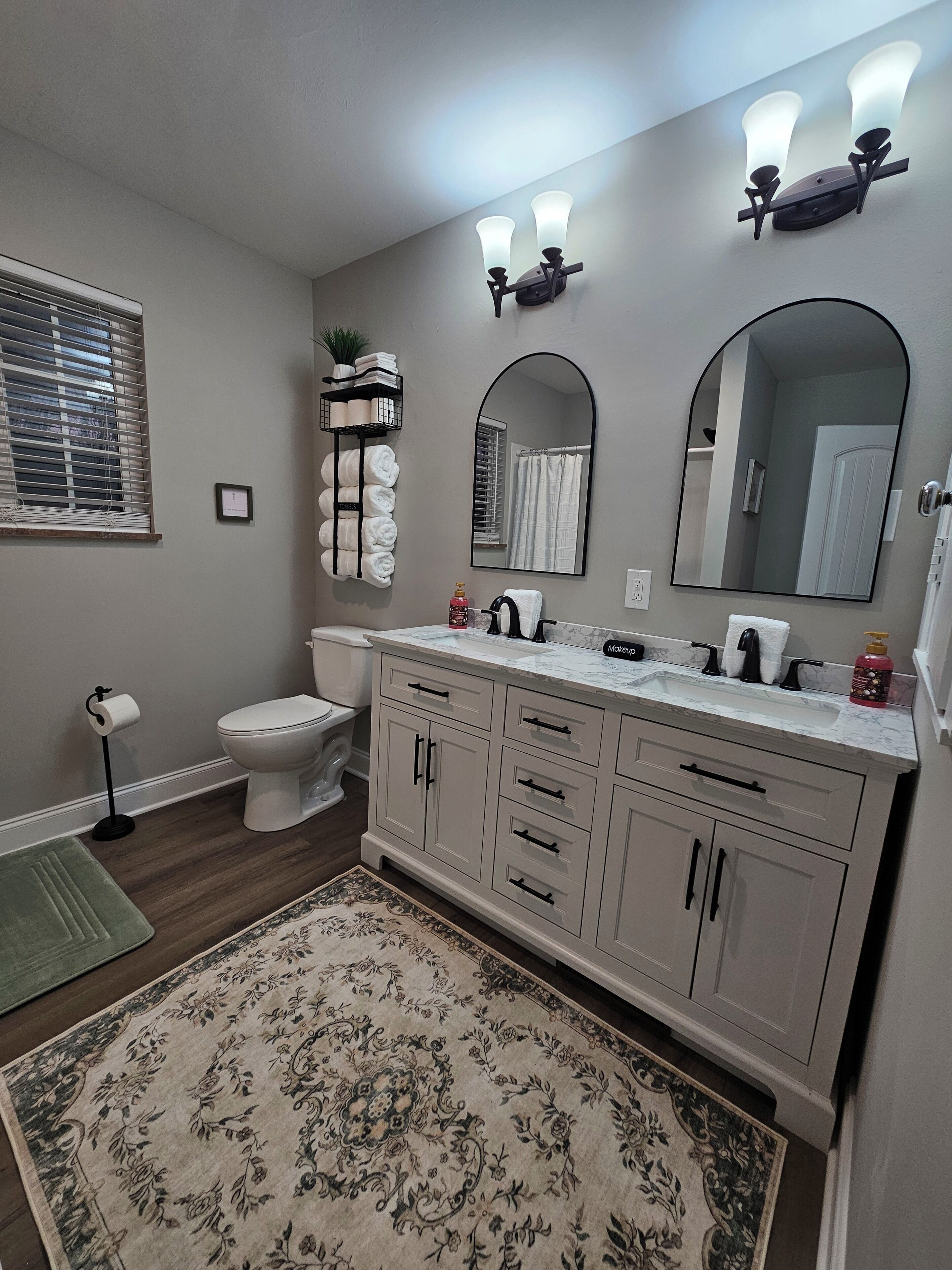 Un baño con inodoro, lavamanos, espejo, toallero y un tapete decorativo.