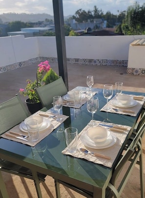 Ein Esstisch mit Geschirr, Gläsern und Besteck, umgeben von Stühlen, auf einer Terrasse mit Blick auf die Landschaft.
