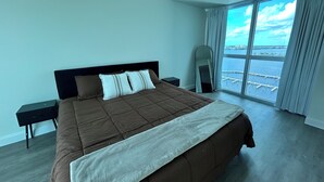 Un dormitorio moderno con una cama grande, una mesita de noche, un espejo y vistas al mar a través de amplias ventanas.