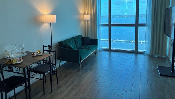 Una habitación con una mesa de comedor, sillas, un sofá verde, dos lámparas de pie y un ventanal con vistas a un puente y al agua.