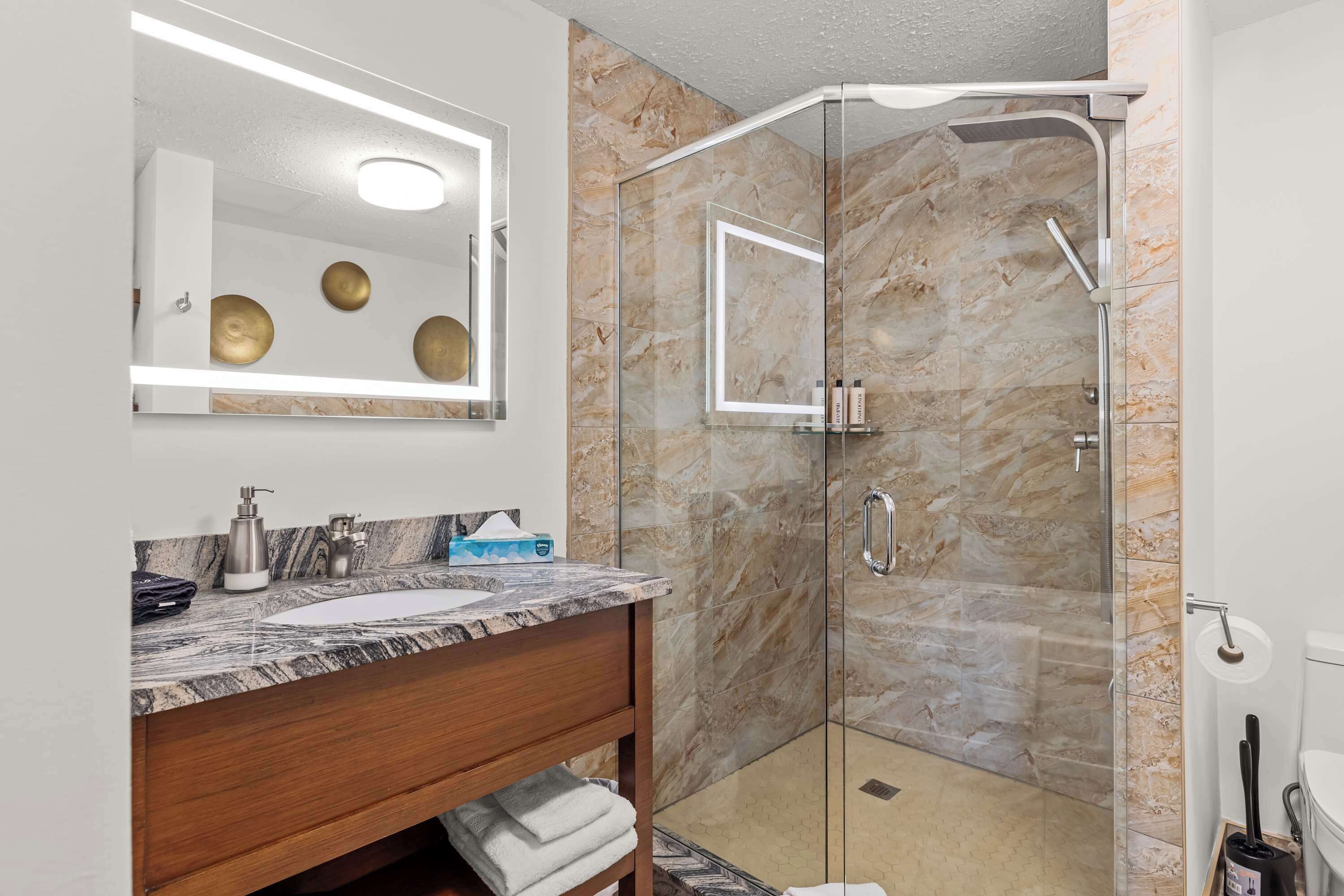Un baño moderno con tocador de mármol, ducha con cerramiento de vidrio y un espejo grande con iluminación.
