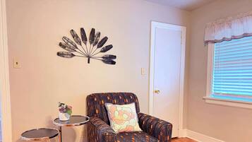 A room with a patterned armchair, a small round table with a vase of flowers, and a decorative wall piece.