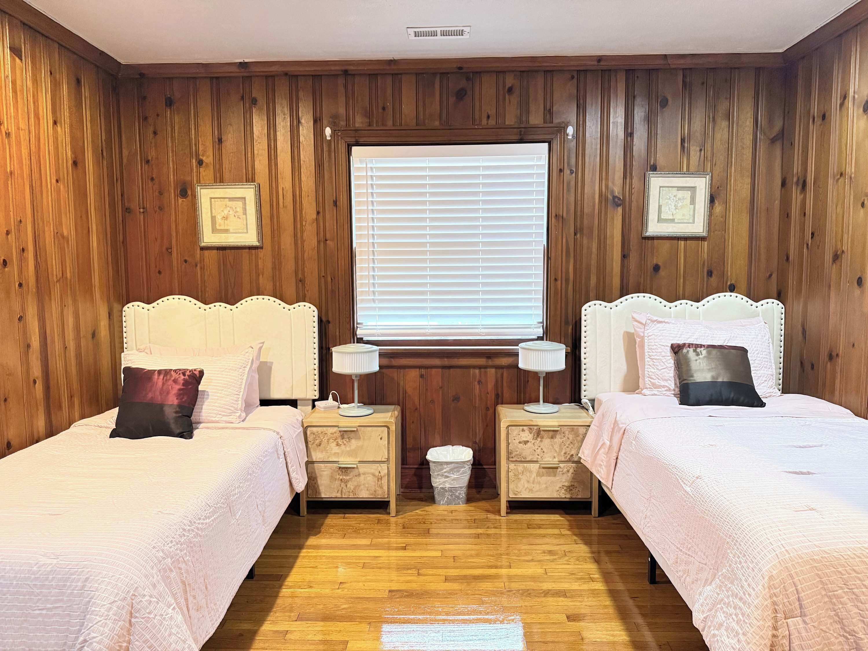 Een kamer met twee bedden, houten wanden, een raam met zonwering en een kleine afvalbak.