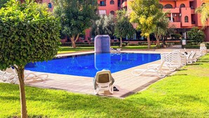 Ein Poolbereich mit einem klaren, blauen Pool, umgeben von grünem Gras und Bäumen. Liegestühle und ein Gebäude mit rosa Wänden sind im Hintergrund zu sehen.