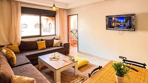 Ein modernes Wohnzimmer mit Sofa, Couchtisch und Esstisch. Ein Fernseher ist an der Wand montiert und ein Fenster mit Vorhängen vorhanden.