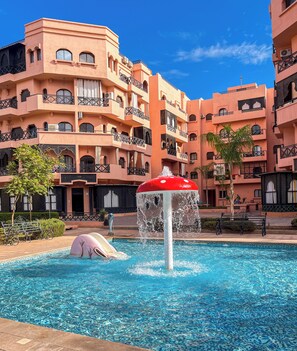 A swimming pool with a water feature, surrounded by multi-story buildings with balconies and a clear blue sky. - Apartment with a view of the pool   (Ouarzazate)