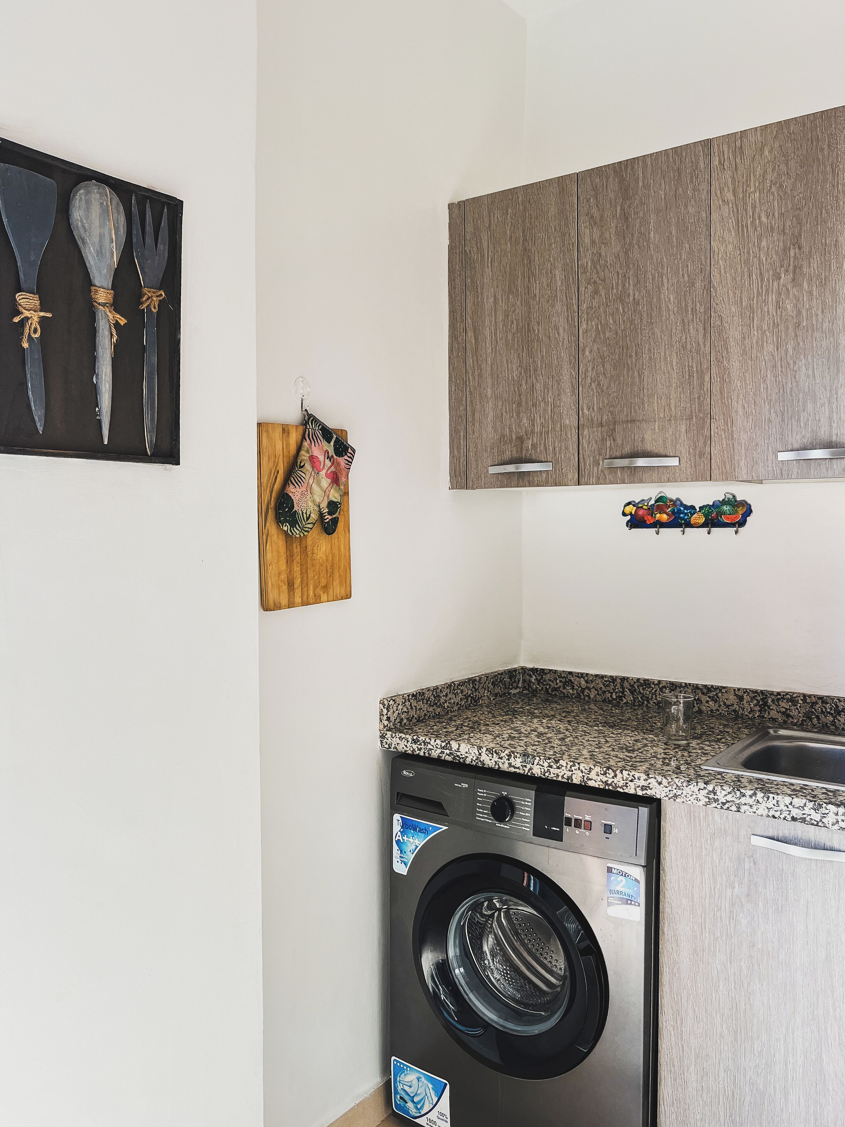 Une cuisine compacte équipée d’un lave-linge, de meubles en bois et d’une décoration murale sur le thème des fruits.
