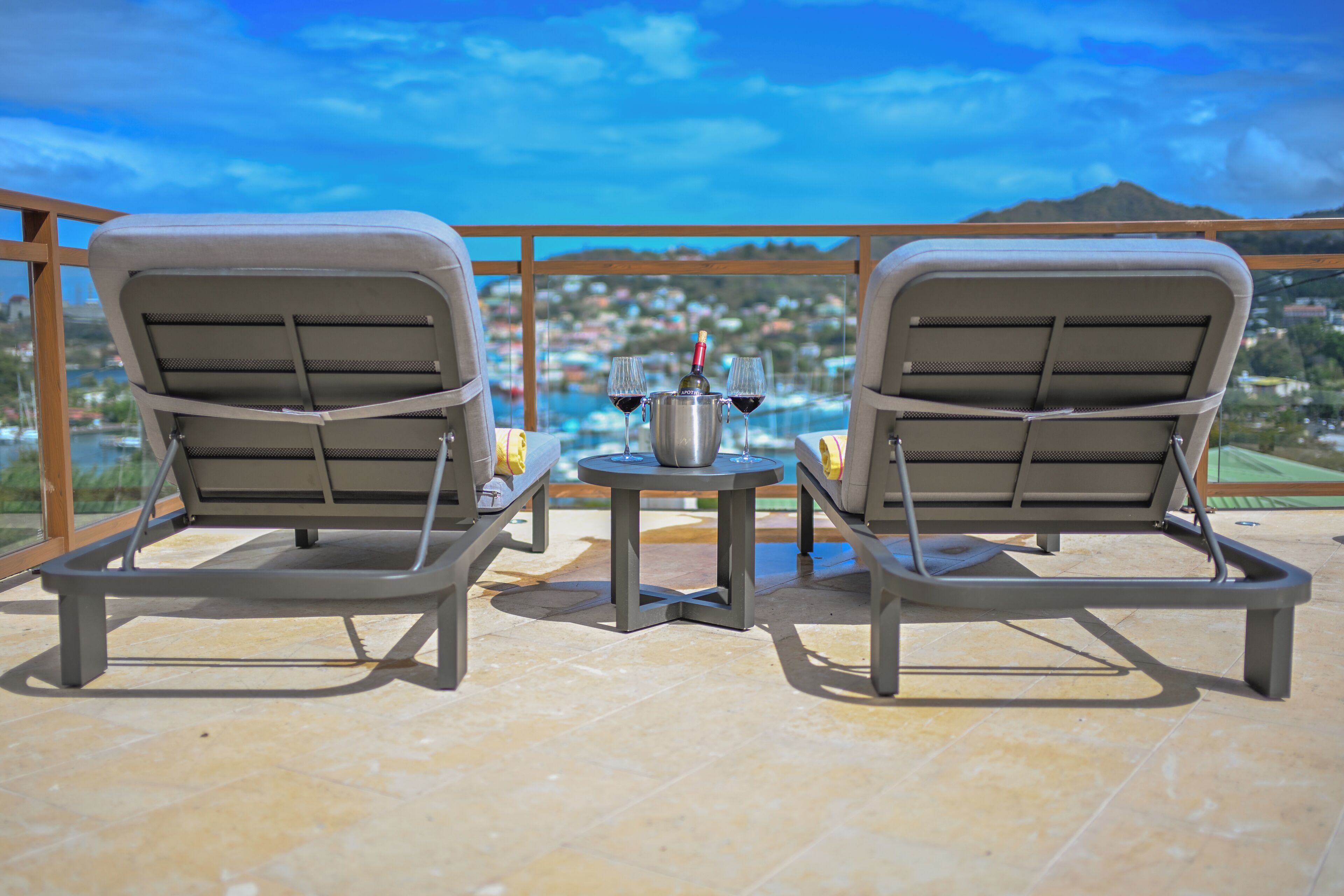 Two lounge chairs facing a pool with a city view, a small table with a bottle and glasses, and a metal grill structure.
