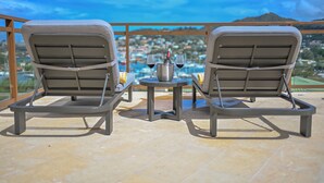 Två solstolar placerade mittemot en pool med utsikt över staden, ett litet bord med en flaska och glas, och en grill av metall.