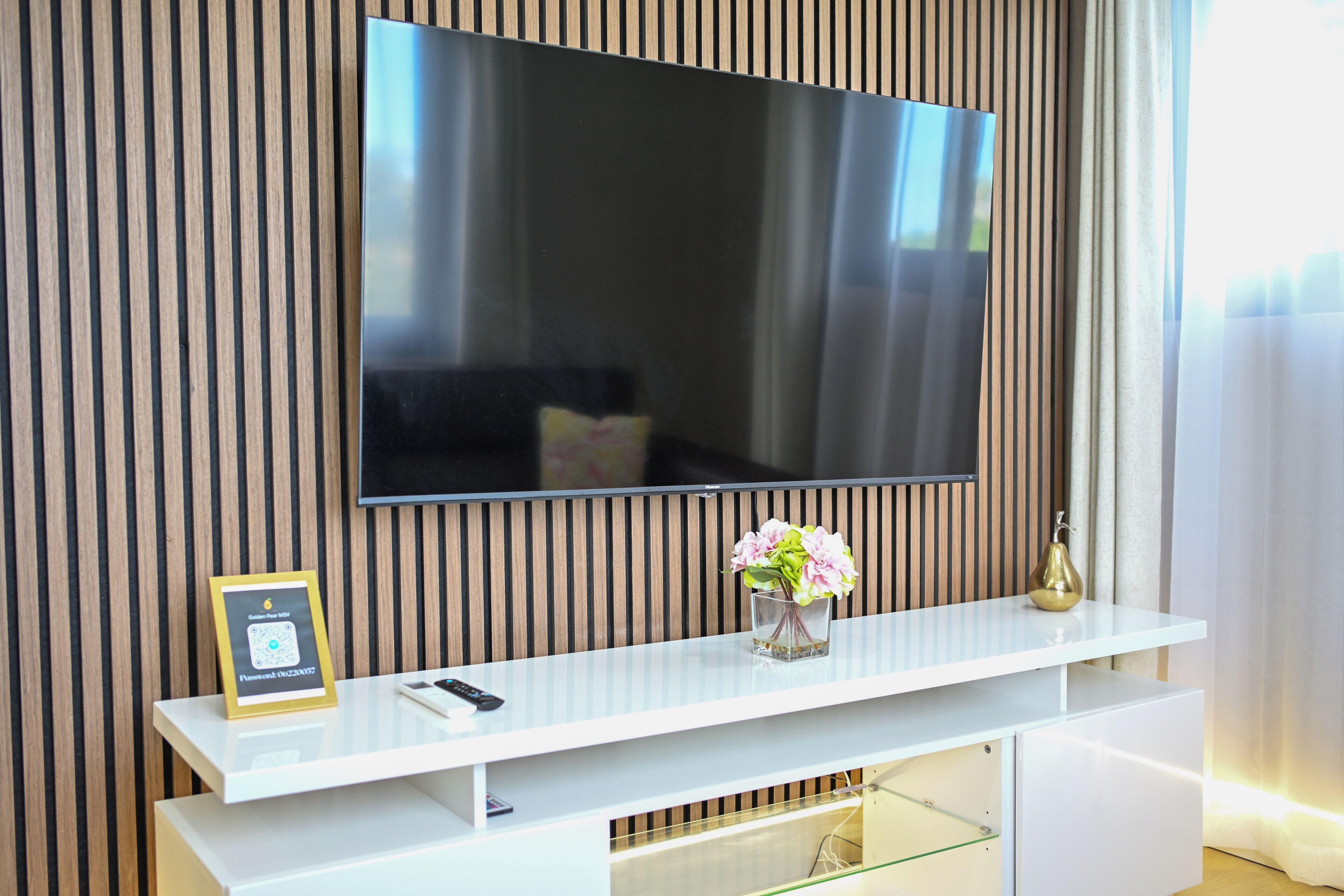 Ett modernt vardagsrum med en platt-TV som är monterad på en väggpanel av trä, ett vitt konsolbord med en vas med blommor och ett inramat certifikat som står på bordet.