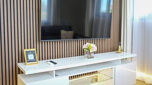 Ett modernt vardagsrum med en platt-TV som är monterad på en väggpanel av trä, ett vitt konsolbord med en vas med blommor och ett inramat certifikat som står på bordet.