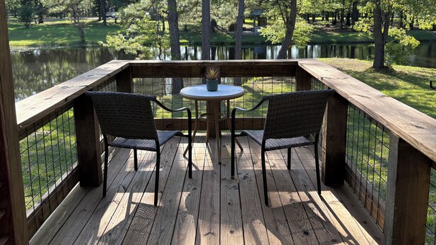 A wooden deck with two wicker chairs and a small round table, overlooking a pond and trees.