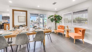 A modern dining area with a large table set for four, surrounded by grey chairs. There is a potted plant and a window with blinds on the wall. - The Loggerhead | Private 3-Unit Oceanview Retreat (Fort Lauderdale)