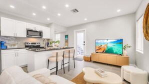 A modern kitchen with a central island, a dining area with bar stools, a living room with a sofa and a coffee table, and a TV displaying a tropical beach scene. - The Loggerhead | Private 3-Unit Oceanview Retreat (Fort Lauderdale)