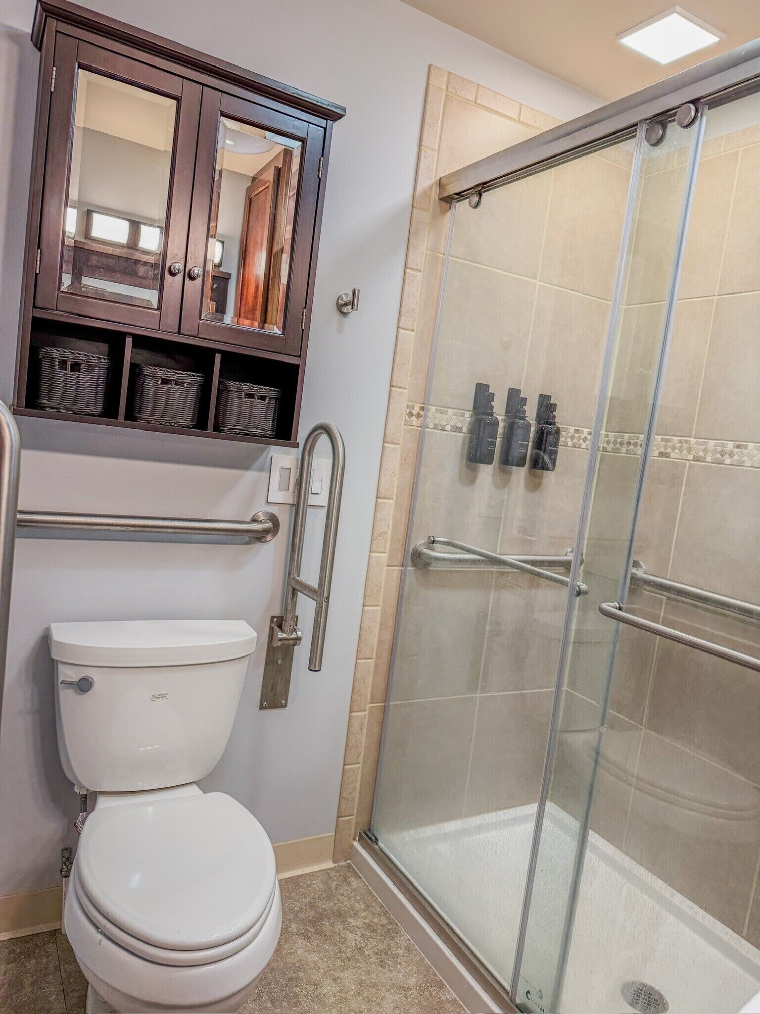Un baño con inodoro, ducha con cerramiento de vidrio, armario de madera con cestas y espejo.