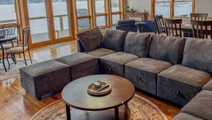 Ein geräumiges Wohnzimmer mit einer großen Eckcouch, einem runden Couchtisch und einem gemusterten Teppich. Große Fenster bieten Blick auf einen See.