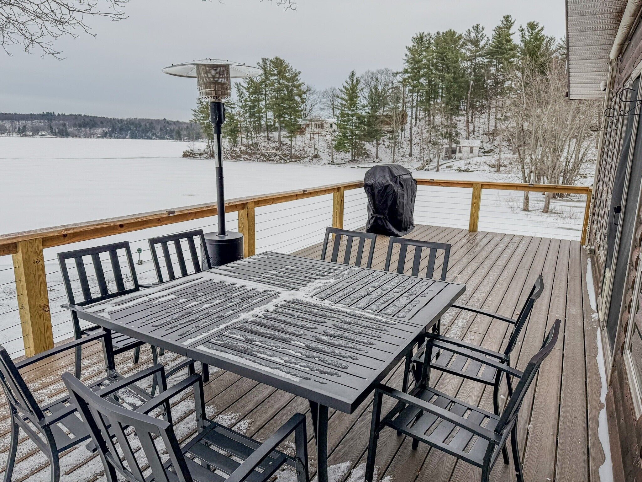 A wooden deck with a metal table and chairs, a covered grill, and a snow-covered landscape in the background.