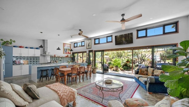 A spacious living area with a dining table, sofas, and a coffee table. There is a kitchen area with cabinets and a countertop. The room has large windows and ceiling fans. - Hyams Reach Retreat - Jervis Bay stay, pets welcome (Shoalhaven City Council)