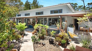 A modern two-story house with a covered patio area, surrounded by various potted plants and a small garden. - Hyams Reach Retreat - Jervis Bay stay, pets welcome (Shoalhaven City Council)