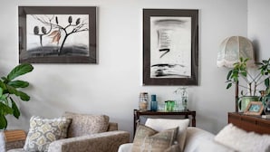 A living room with two framed abstract paintings, a sofa with patterned cushions, a side table with decorative items, and a potted plant. - Hyams Reach Retreat - Jervis Bay stay, pets welcome (Shoalhaven City Council)