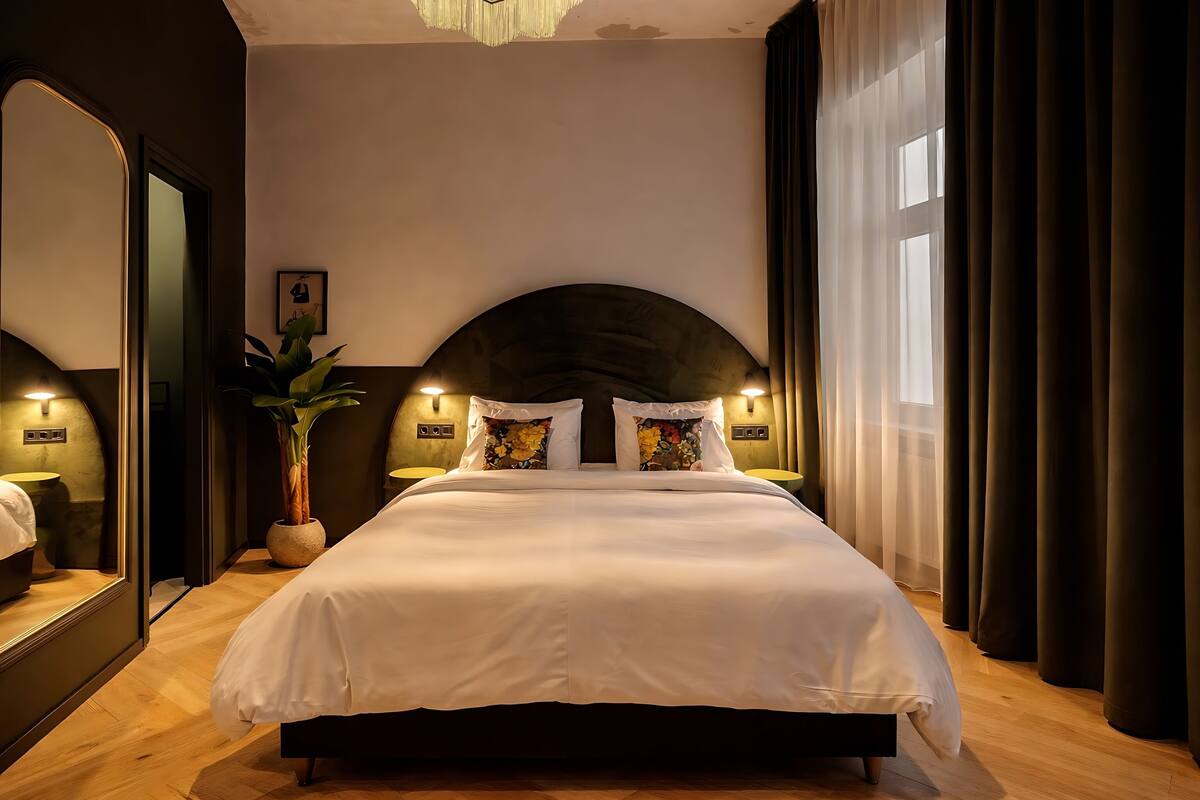 Un grand lit avec une tête de lit sombre, une plante en pot et un miroir sur un des murs.