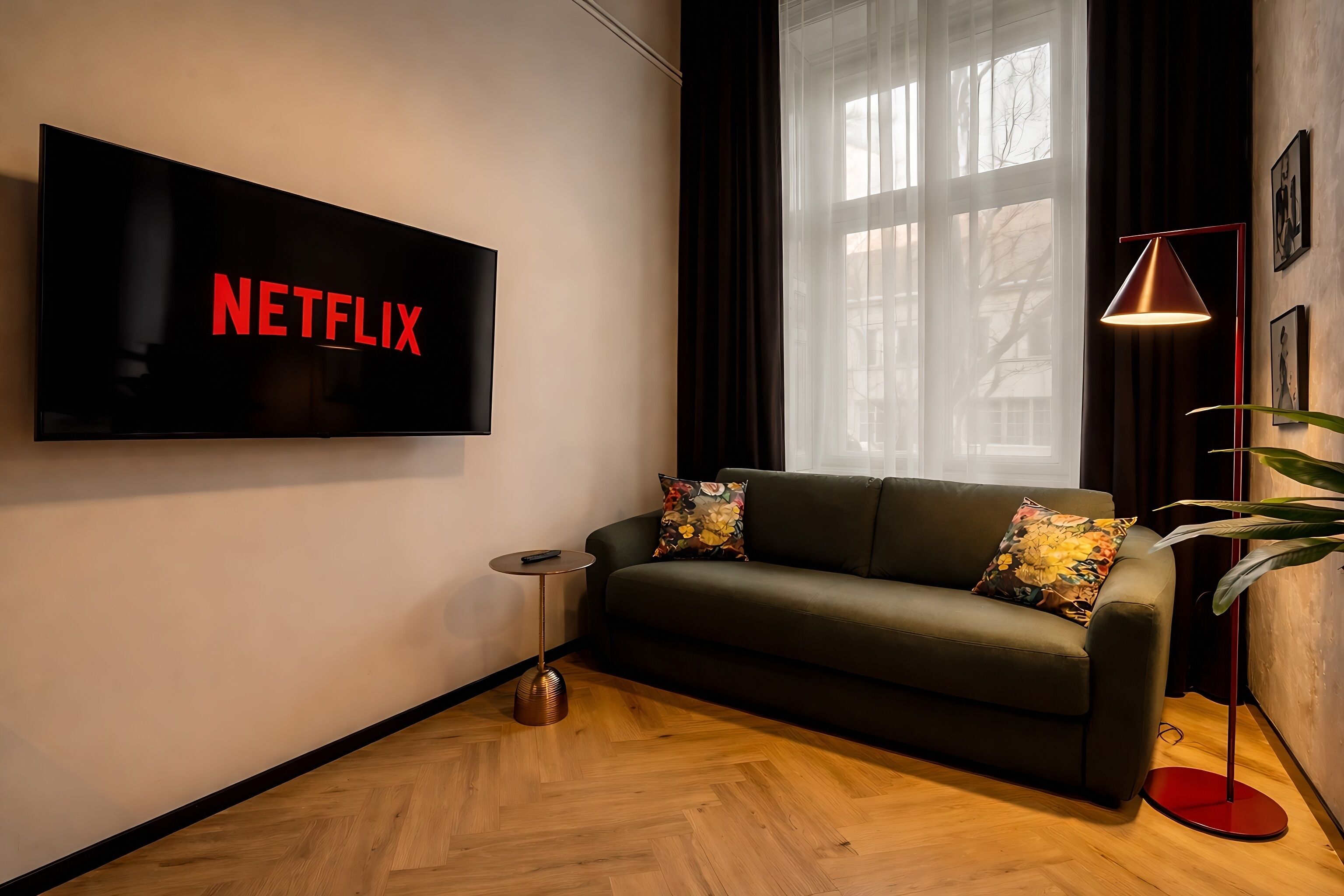 Un salon moderne doté d’un grand téléviseur à écran plat affichant le logo de Netflix, d’un canapé vert foncé agrémenté de coussins à motifs floraux, d’une petite table d’appoint, d’un lampadaire et d’une grande fenêtre ornée de rideaux légers.