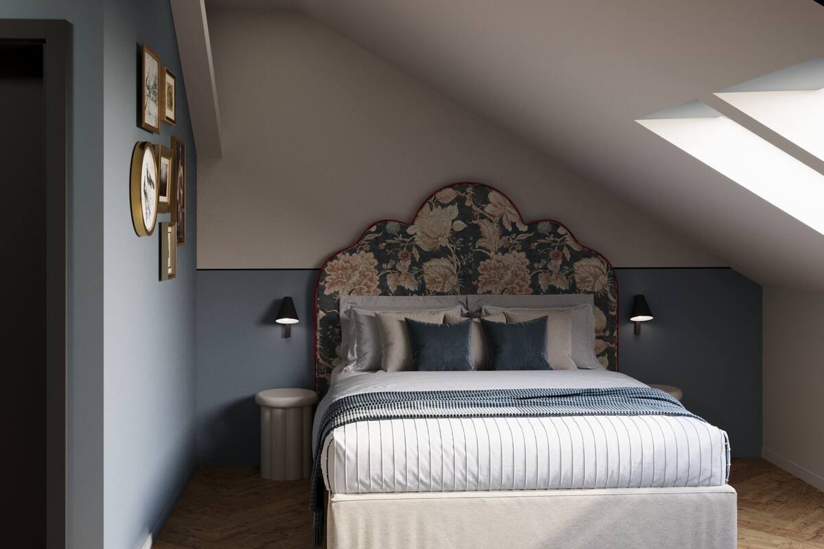 Une chambre avec un plafond en pente, un lit doté d’une tête de lit à motifs floraux, deux lampes fixées au mur et un petit tabouret rond.