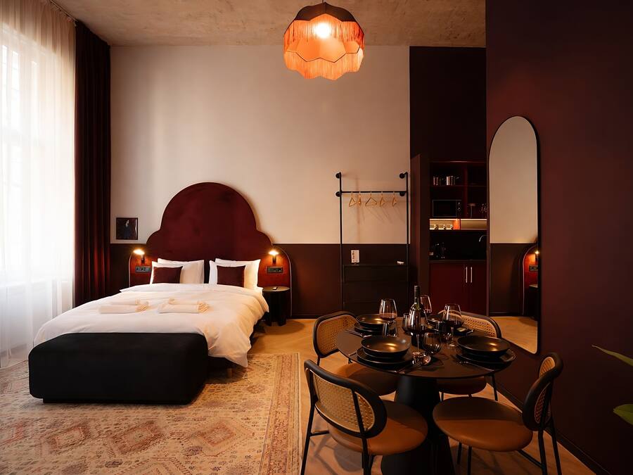 A hotel room with a bed, bedside table, lamp, and a dining area with chairs and a table.