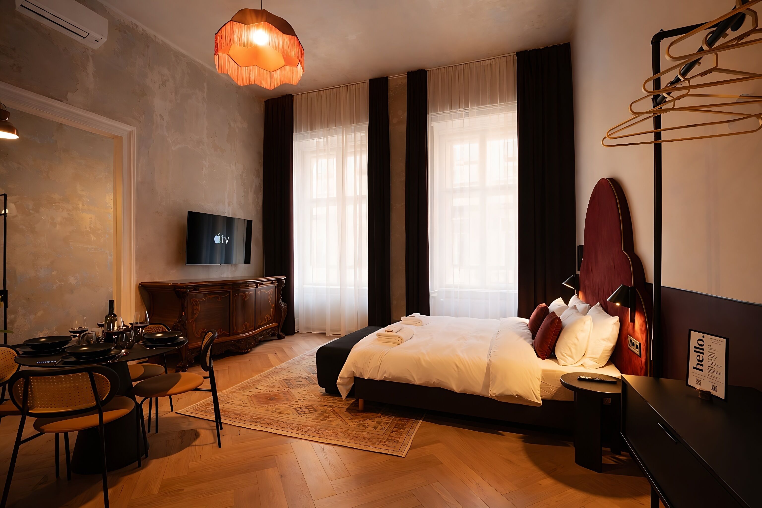 A hotel room with a bed, a desk with a flat-screen TV, a guitar, and a dining area with chairs and a table.