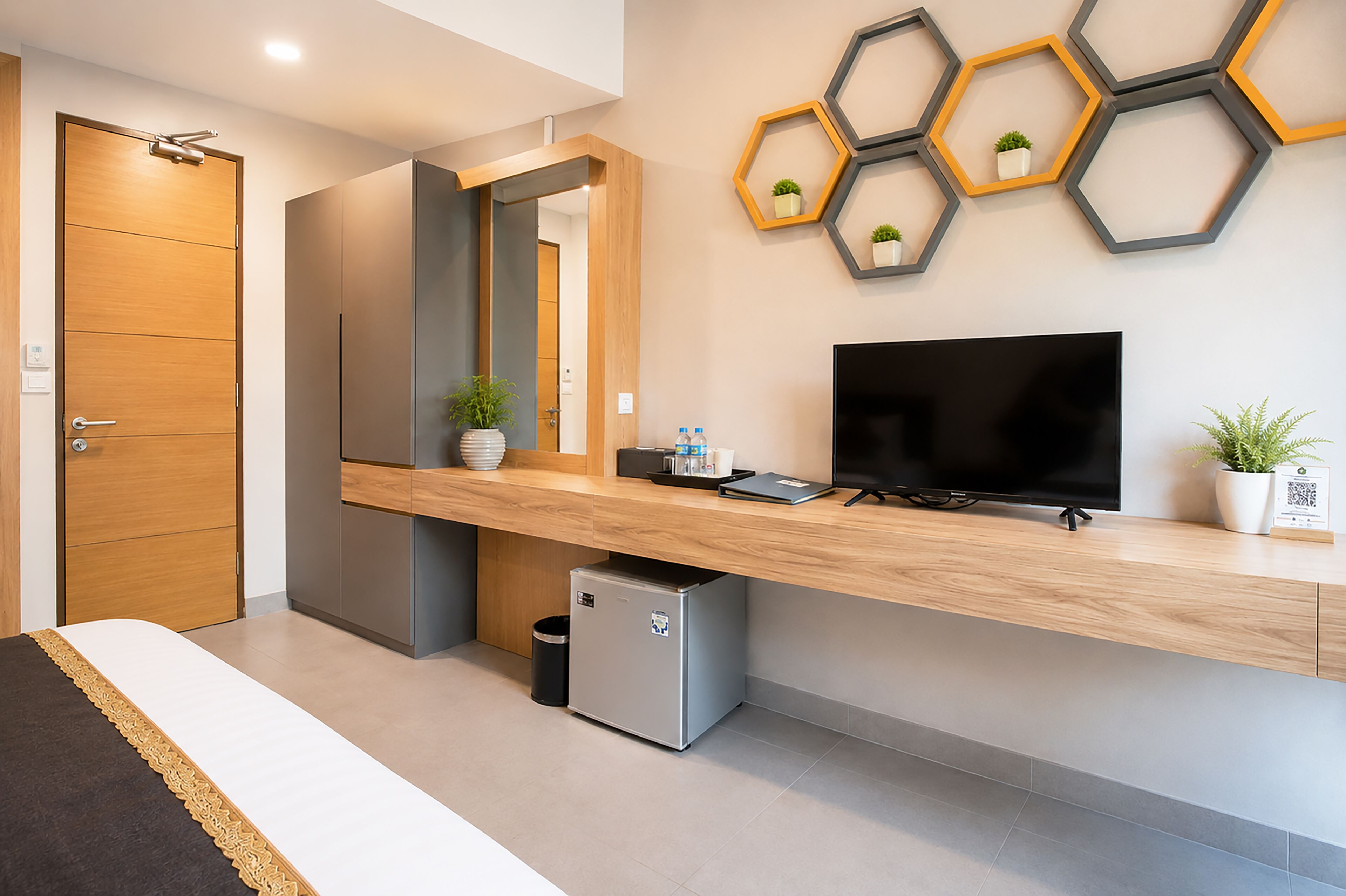 A modern hotel room with a wooden desk, a flat-screen TV, a small refrigerator, and a mirror.