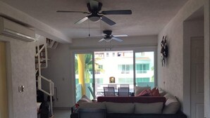 A living room with a sofa, a dining table, and a ceiling fan. There is a staircase on the left and a view of a building outside. - APARTMENT IN ACAPULCO DIAMOND ZONE (Acapulco de Juárez)