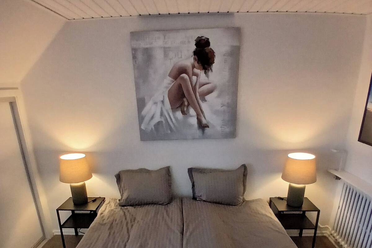 A bedroom with a large bed, two bedside tables with lamps, a wall-mounted TV, and a painting above the bed.