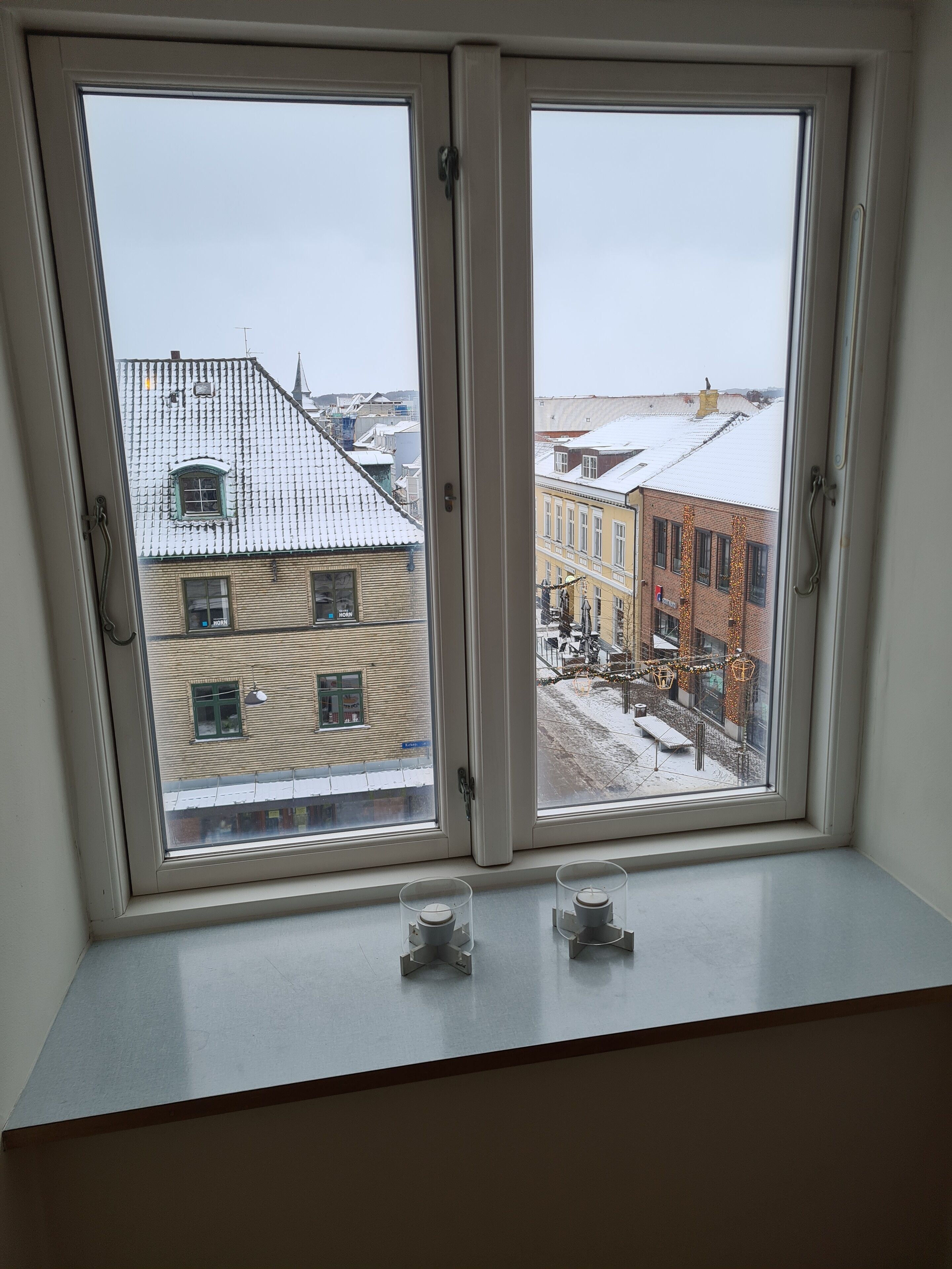 A window with two panes, overlooking a street with buildings and a snow-covered ground.