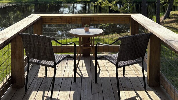 A wooden deck with two wicker chairs and a small round table, overlooking a pond and surrounded by trees.
