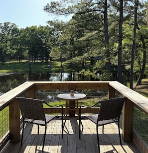 A wooden deck with two wicker chairs and a small round table, overlooking a pond and surrounded by trees.