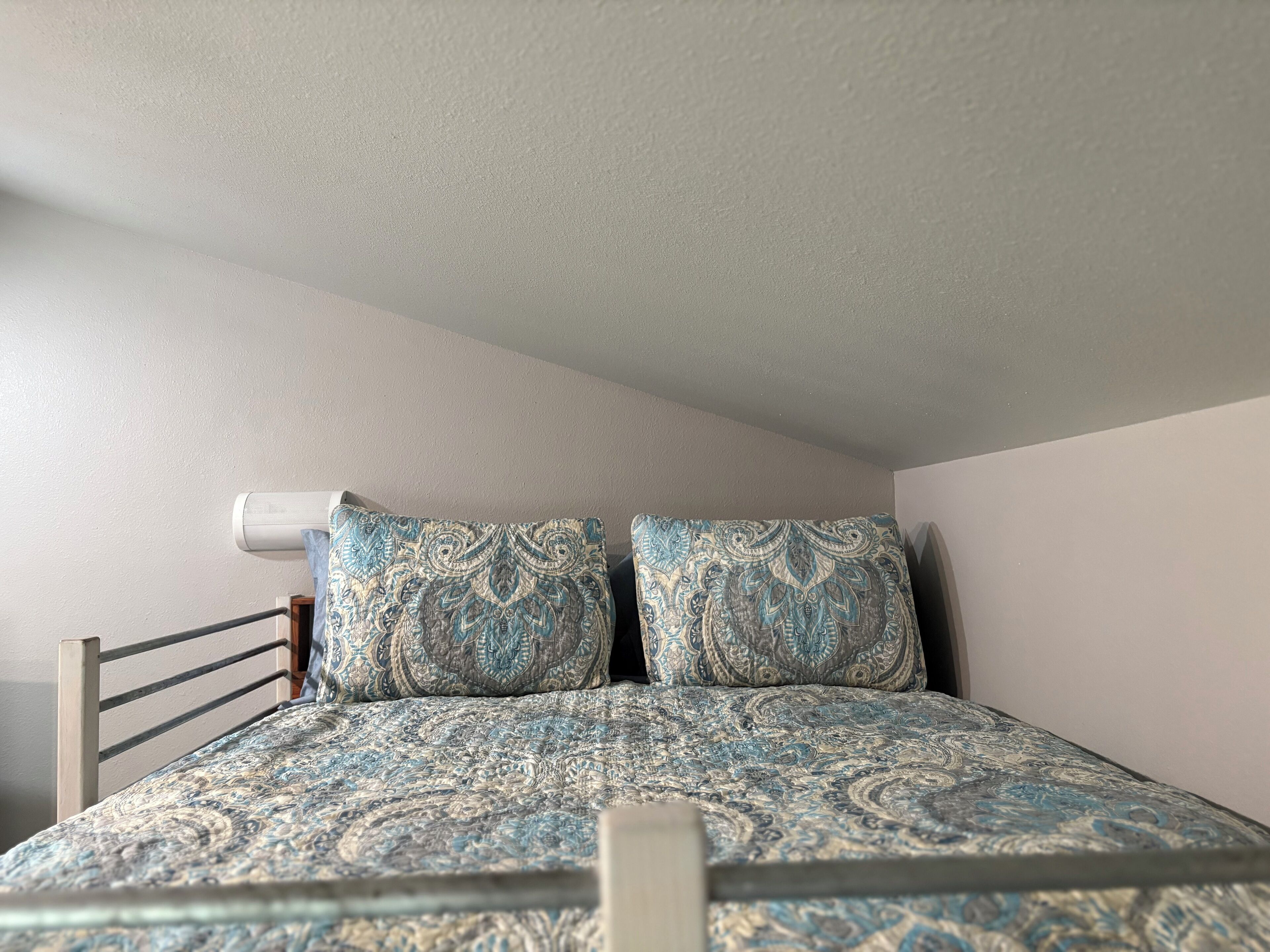 A bedroom with a bed, two patterned pillows, a headboard, and a wall-mounted air conditioner.