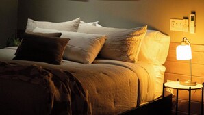 Un dormitorio con poca luz, una cama, almohadas, una mesita de noche con una lámpara y dos cuadros en la pared.