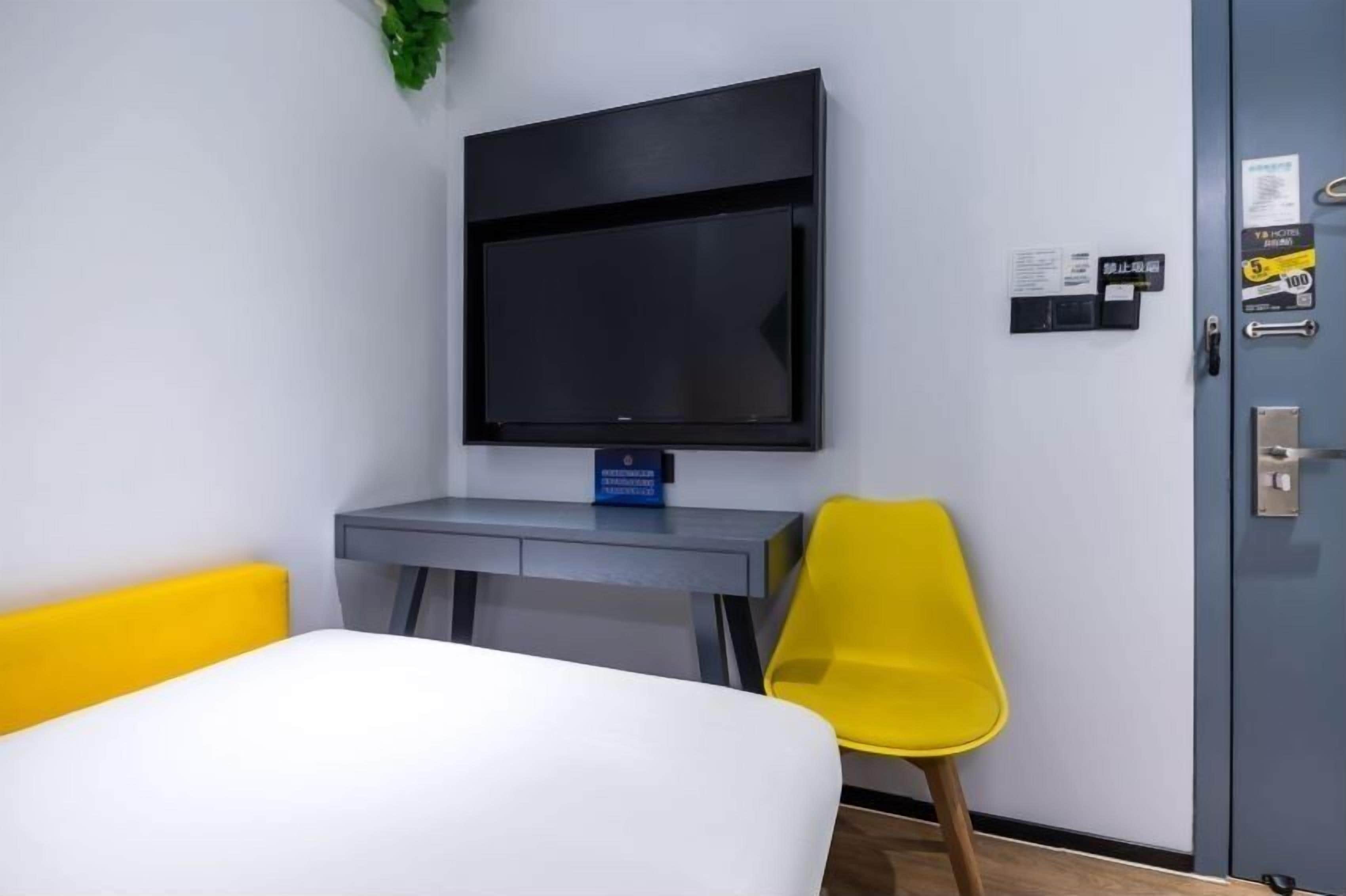 A hotel room with a bed, a desk, a television mounted on the wall, a yellow chair, and a door with a lock and a key.