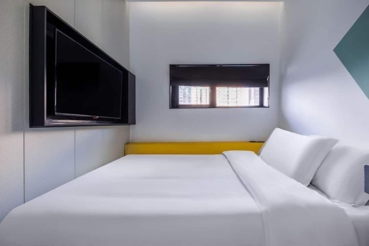 Une chambre d’hôtel avec un lit, un téléviseur fixé au mur, une fenêtre avec des stores et un mur d’accent jaune.