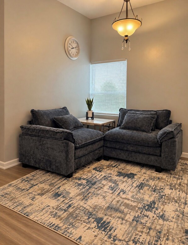 A living room with two grey sofas, a small table, a clock, and a window with blinds. - Cozy 2BR Downtown Retreat Just 5 Min to UF Health (Jacksonville)