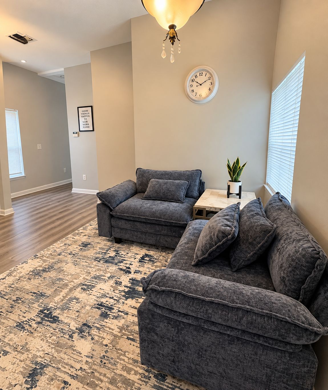 A living room with two grey sofas, a patterned rug, a small table with a plant, a wall clock, and a motivational quote sign.