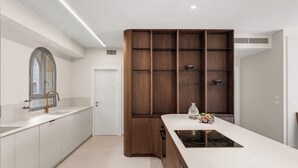 A modern kitchen with white cabinetry, a built-in wooden shelving unit, a central island with a stove, and a window with a curved mirror. - 3-bedroom house in good condition with cool AC (Woodmere)
