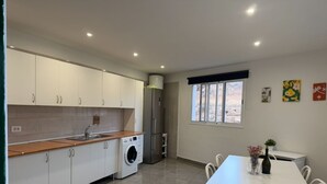 Una cocina moderna con armarios blancos, lavadora integrada, comedor con mesa y sillas blancas, y una ventana decorada con una obra de arte.