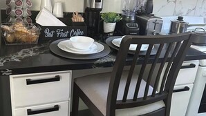 Eine moderne Küche mit weißen Schränken, einer schwarzen Arbeitsplatte und einem dunklen Holzhocker. Es gibt eine Kaffeestation mit einem Schild, einem Mixer und einem Toaster.