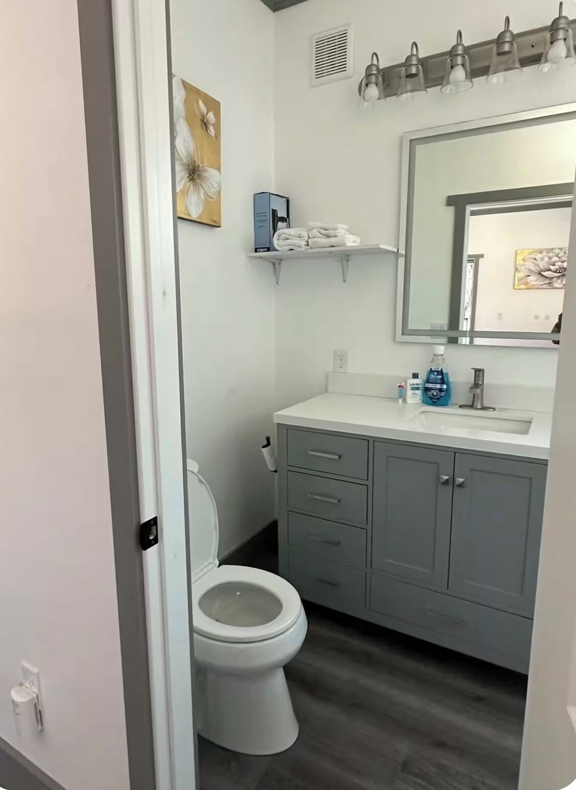 Un bagno con il water, un lavabo e uno specchio. Ci sono mensole sopra il lavabo e un quadro al muro.