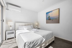 A hotel room with a bed, two bedside tables, a wall-mounted air conditioner, and a beach scene painting.