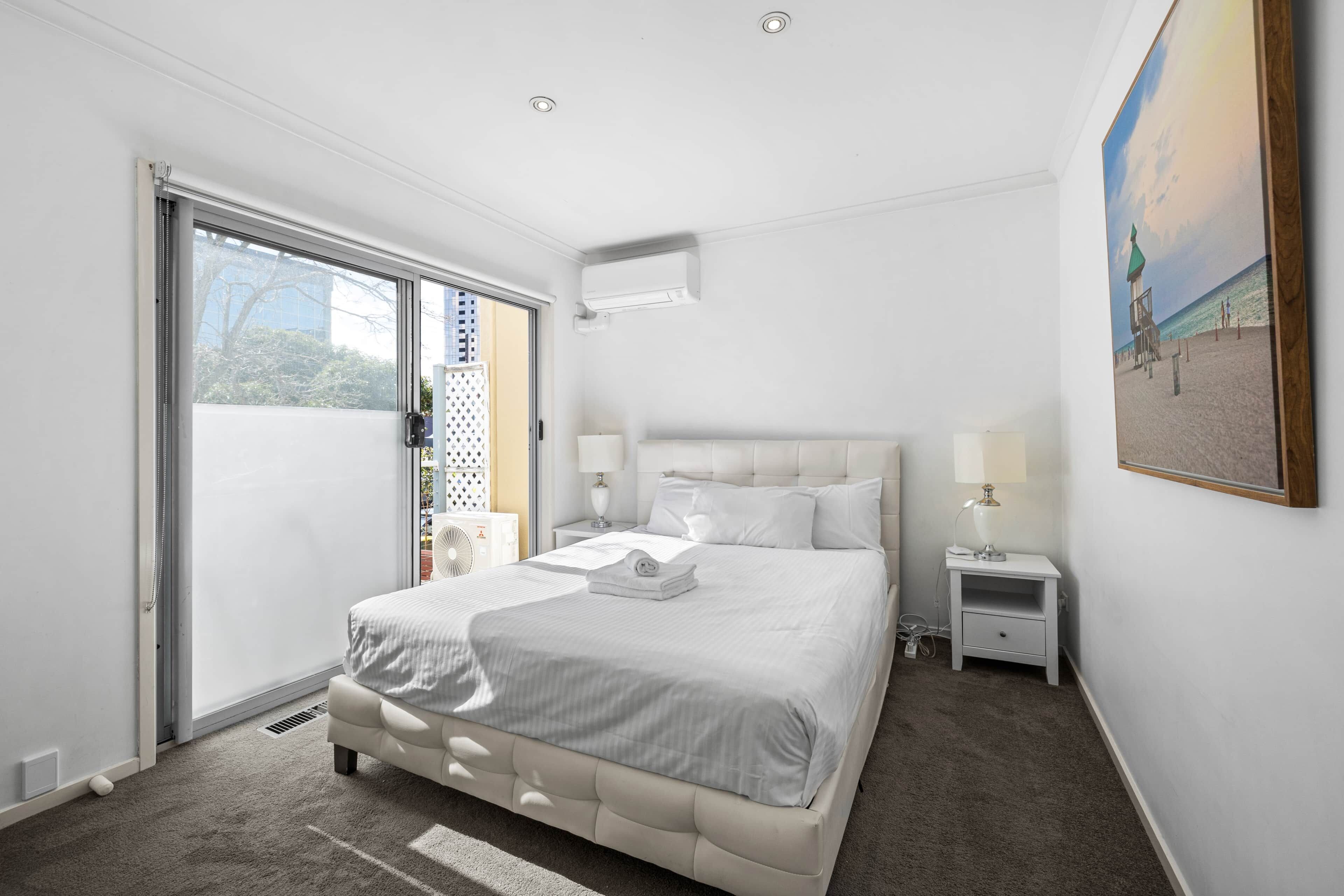 A hotel room with a large bed, a bedside table with a lamp, a window with a view of the outside, and an air conditioning unit on the wall.