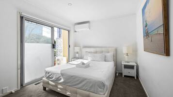 A hotel room with a large bed, a bedside table with a lamp, a window with a view of the outside, and an air conditioning unit on the wall.
