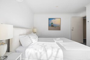 A hotel room with a bed, a nightstand, a lamp, a painting of a beach scene, and a door. - Sleep 5 | Luxury Box Hill Townhouse (Box Hill)
